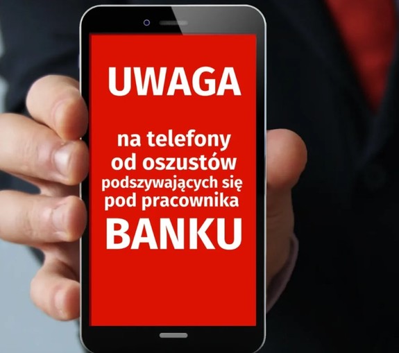 Telefon komórkowy trzymany w dłoni z apisem na ekranie &amp;quot;Uwaga na telefony od oszustów podających się za pracownika banku&amp;quot;.