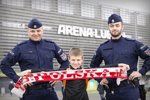 Policjanci z chłopcem w tle stadion. Chłopiec trzyma szalik z napisem Polska.