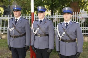 Trzech policjantów stoi na baczność pod masztem z flagą Polski.