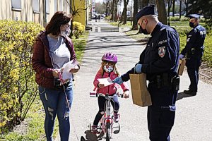 Policjant umundurowany przekazuję dziecku na rowerku maseczkę ochronną obok stoi mama dziewczynki.