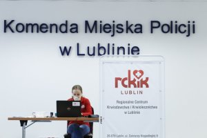 Pracownik Regionalnego Centrum Krwiodawstwa i Krwiolecznictwa w Lublinie siedzi przy laptopie. Za nią duży napis Komenda Miejska Policji w Lublinie.