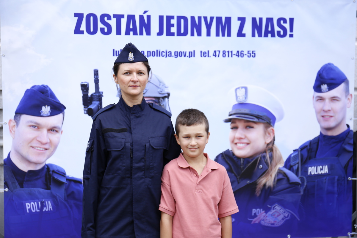 Wspólne zdjęcie nowej policjantki z chłopczykiem na tle baneru zachęcającego do wstąpienia w szeregi. policji.