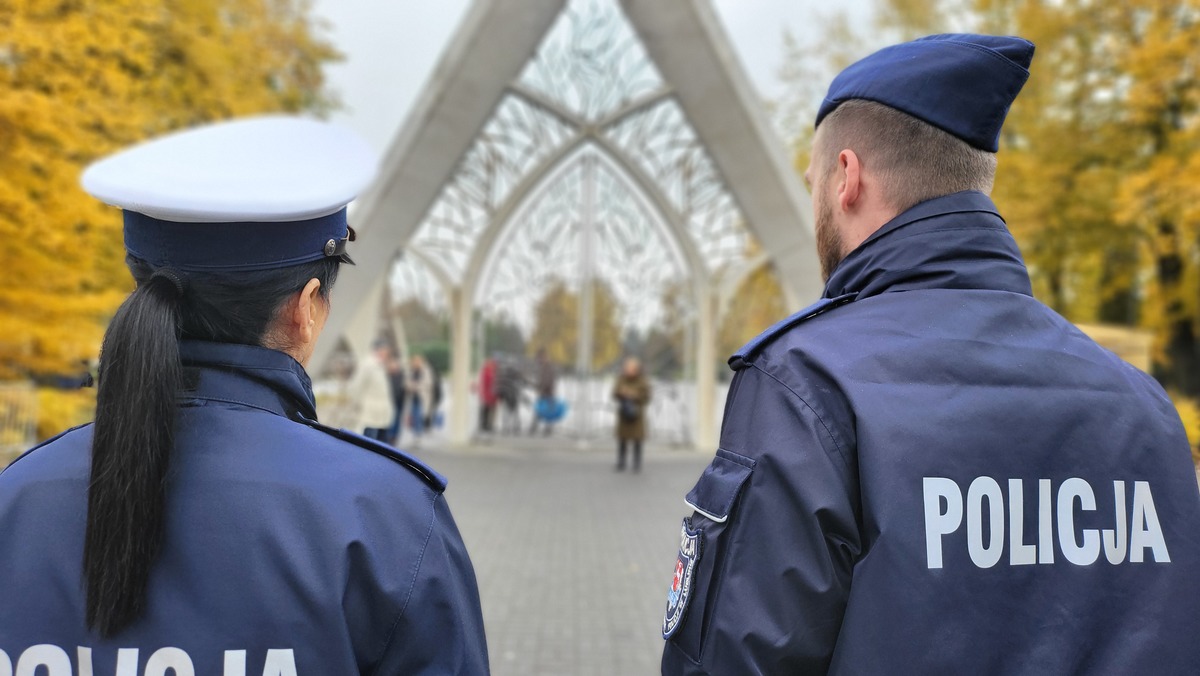 Policjanci w pierwszym planie zdjęcia w głębi widać bramę do cmentarza.