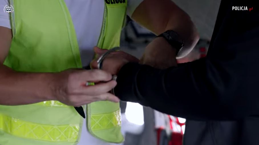Kadr z filmu na którym widać jak policjant zakłada kajdanki na ręce zatrzymanemu mężczyźnie..