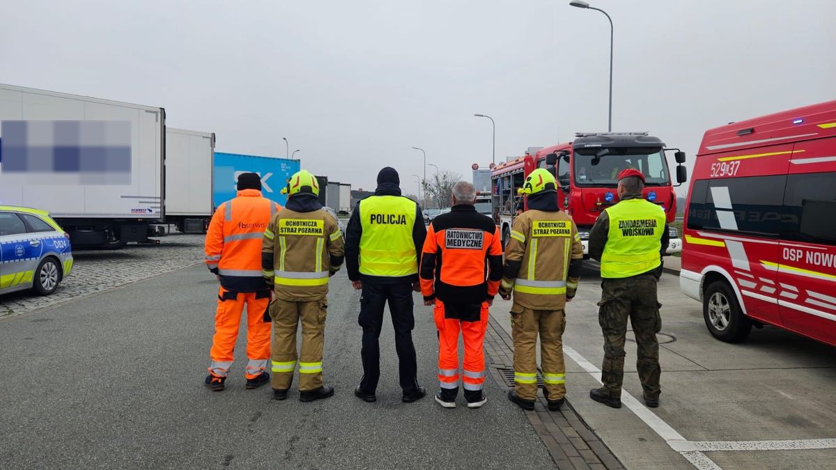umundurowani funkcjonariusze służb stoją tyłem. W tele znajdują się samochody ciężarowe, radiowóz policyjny i wozy strażackie