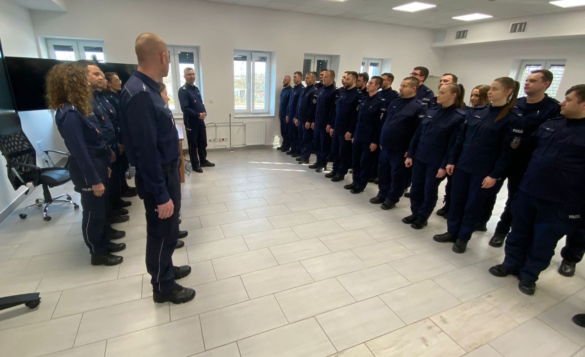 Uroczysta zbiórka policjantów na sali dydaktycznej. Policjanci ustawieni w szeregu są zwróceni frontem do kadry dydaktycznej 