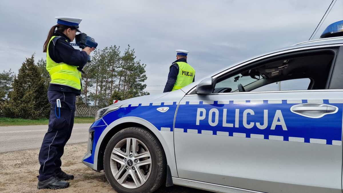 Policjantka ruchu drogowego stoi przy radiowozie, mierzy prędkość trzymając w dłoniach ręczny miernik prędkości