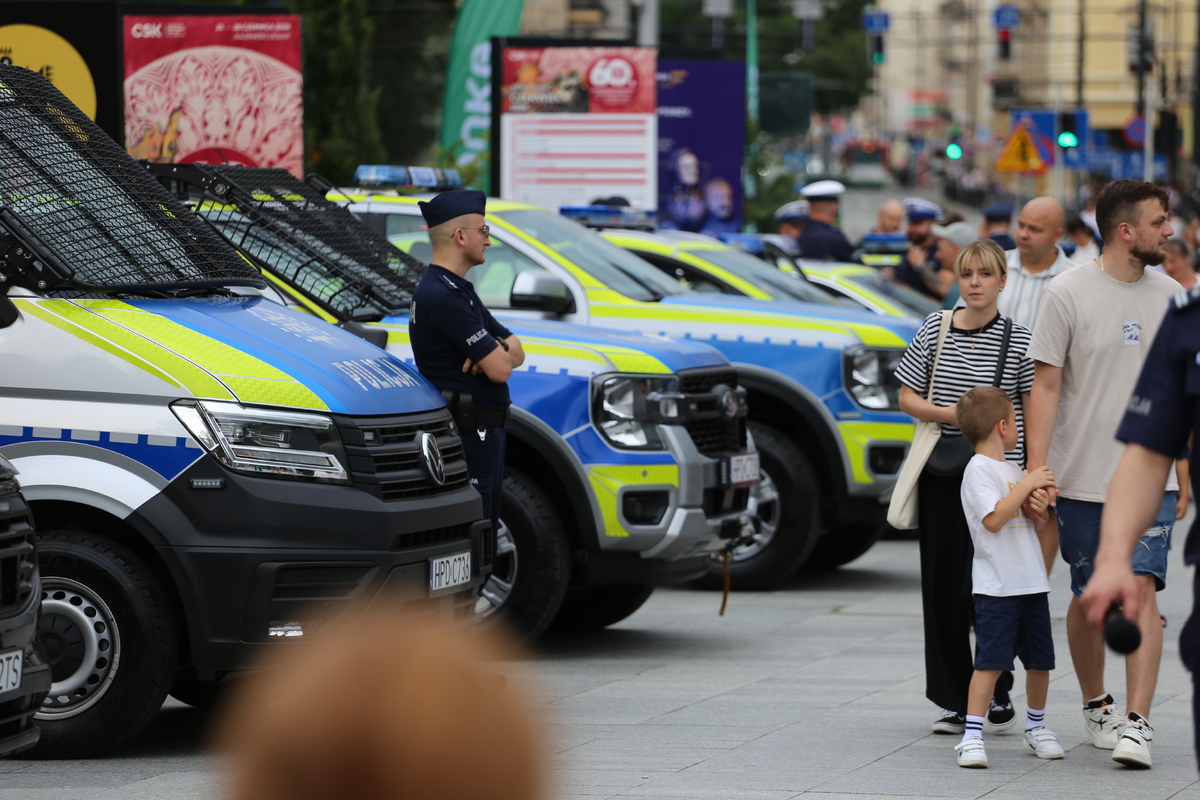 policjant na tle ustawionych w rzędzie radiowozów. W tle uczestnicy pikniku 