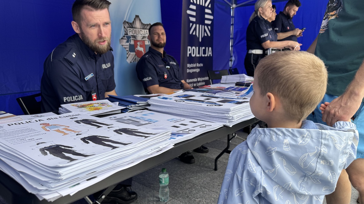 pod policyjnym namiotem policjanci, na stole ulotki, małe dziecko przy stoisku 