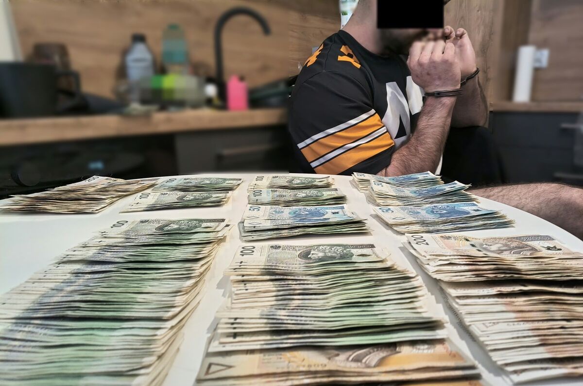 na stole ułożone banknoty polskich złotych, w tle siedzący na krześle zatrzymany mężczyzna z rękami zakutymi w kajdanki