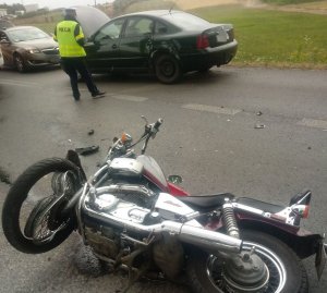 Na pierwszym planie leżący na jezdni motocykl z uszkodzonym przednim kołem, a dalej samochód volkswagen i stojący przy nim umundurowany policjant
