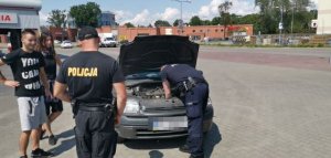 policjant naprawia uszkodzony samochód, obok którego stoi dwójka uczestników festiwalu