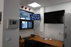 Pomieszczenie dyżurnego komisariatu. Na ścianie umieszczone są dwa monitory z podglądem na otoczenie i pomieszczenia nowej siedziby. Na biurku leżą telefon, lampa oraz komputer.