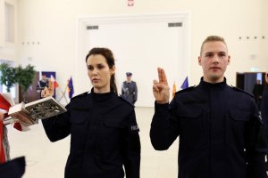 Policjant trzyma sztandar Komendy Wojewódzkiego Policji w Lublinie. Nowi policjanci ślubują z uniesioną prawą ręką do góry z wyprostowanymi dwoma palcami.
