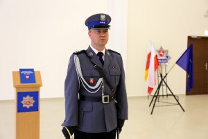 Dowódca uroczystości  ubrany w mundur galowy koloru granatowego w ręku trzyma szablę. Za policjantem na mównicy leży Konstytucja Rzeczypospolitej Polskiej.