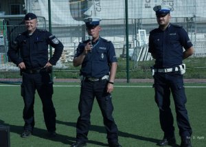 Trzech policjantów stojących na boisku, jeden trzyma mikrofon