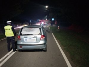 miejsce wypadku drogowego, umundurowany policjant stoi na drodze, przed nim samochód marki Hyundai, w tle pojazd bojowy straży pożarnej