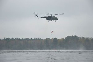 Unoszący się nad taflą jeziora wojskowy śmigłowiec, a pod nim na linie wciągany z wody do góry ratownik