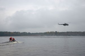 Unoszący się nad taflą jeziora wojskowy śmigłowiec, po prawej na wodzie łódź motorowa z ratownikami