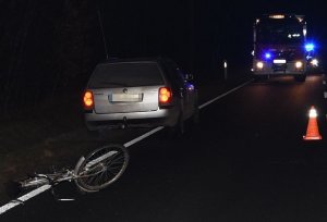 leżący na jezdni uszkodzony rower i stojący w pobliżu samochód marki Volkswagen Passat, w tle samochód Straży Pożarnej i Policji