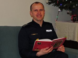 Policjant siedzi na kanapie, w ręku trzyma książkę, w tle dekoracje świąteczne.