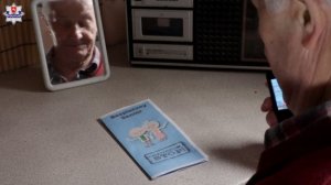 starszy mężczyzna z telefonem przy uchu, w tle odbijająca się w lusterku jego twarz oraz leżąca na stole niebieska kartka z namalowanymi postaciami dwóch osób oraz napisem bezpieczny senior, oszustom stop