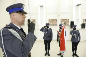 Na zdjęciu policjant trzyma sztandar w poczcie sztandarowym. Po lewej stronie zdjęcia prowadzący uroczystość oddaje honor trzymając szable.
