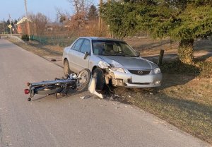 Leżący na jezdni uszkodzony motocykl marki MZ. Po prawej stronie na poboczu jezdni pojazd marki Mazda z uszkodzonym przodem z prawej strony.