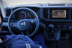 Kokpit radiowozu marki Volkswagen Crafter, gdzie widzimy kierowcę policjanta oraz kierownicę, monitor czy drążek automatycznej zmiany biegów.