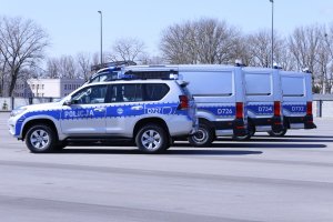 Toyota Land Cruiser i Volkswageny Craftery na placu obok siebie. Radiowozy są koloru srebrno niebieskiego z napisem policja.