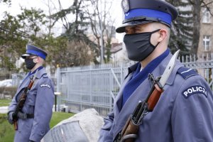 Dwóch policjantów z bronią długą w maseczkach zasłaniających usta i nos stoi po obu stronach pomnika.
