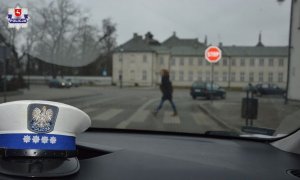 Radiowóz drogówki stoi przed przejściem dla pieszych. Kobieta idzie po pasach. W tle policyjna czapka na kokpicie.