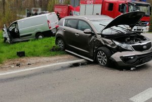 Pojazd marki KIA z rozbitym przodem na środku jezdni. Na dalszym planie  poza drogą pojazd maki Peugeot Expert oraz wóz strażacki.