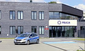 W pierwszym planie zdjęcia radiowóz oznakowany policji. W drugim planie zdjęcia budynek pierwszego komisariatu policji w Lublinie. Budynek ma kolor szary. Nad wejściem napis policja i logo policji.