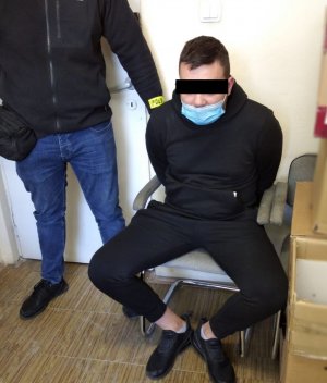 Na krześle siedzi zatrzymany 22-latek. Obok stoi policjant po cywilnemu