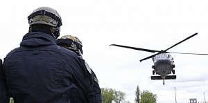 Po lewej stronie zdjęcia policjanci w kombinezonach i kaskach ochronnych obserwują lecący Black Hawk.