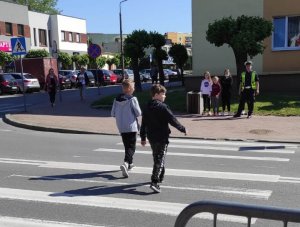 dwaj chłopcy przechodzą przez przejście dla pieszych, po drugiej stronie drogi stoi umundurowany policjant