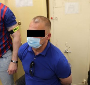 Zatrzymany mężczyzna siedzi na krześle. Obok stoi policjant kryminalny