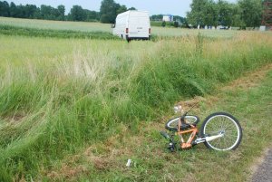 leżący rower, na poboczu trawiastym, w oddali samochód VW  w polach