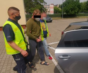 mężczyzna w kajdankach zespolonych w asyście policjantów podchodzi do samochodu