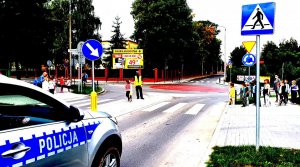 policjant uczy dzieci jak prawidłowo przechodzić przez jezdnię, na zdjęciu widać również radiowóz
