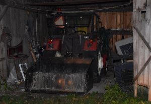 zdjęcie w nocy. ciągnik rolniczy z podczepioną czarną skrzynią stoi wewnątrz drewnianego pomieszczenia gospodarczego