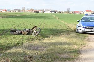 leżący na polu uszkodzony motocykl, stojący obok na drodze radiowóz