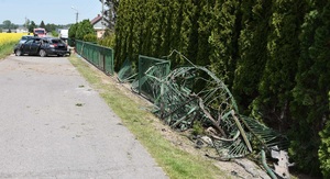 Uszkodzone metalowe ogrodzenie po prawej stronie drogi. Na jezdni stoi uszkodzony samochód.