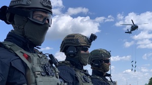 Trzech kontrterrorystów w pierwszym planie zdjęcia za nimi policyjny śmigłowiec z podwieszonymi na linach funkcjonariuszami.