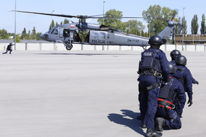W pierwszym planie po prawej stronie zdjęcia policjanci ubrani w umundurowanie bojowe. W drugim planie zdjęcia lądujący śmigłowiec Black Hawk.