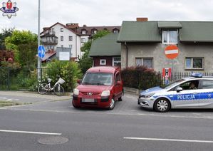 stojący na skrzyżowaniu ulic samochód osobowy marki Peugeot i policyjny radiowóz. Oparty o ogrodzenie posesji  rower