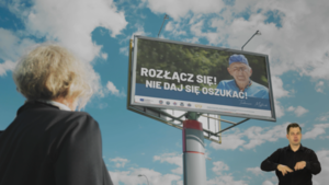 Zdjęcie przedstawia starszą kobietę patrzącą w bilbord na którym jest inny starszy mężczyzna i napis "Rozłącz się nie daj się oszukać"