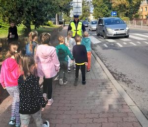 policjant stoi przy przejściu dla pieszych. w stronę przejścia zmierza grupa dzieci. widać oczekujący przed przekściem