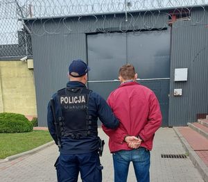 Zdjęcie na którym widoczna jest osoba poszukiwana trzymana przez umundurowanego policjanta, w tle widać bramę zakładu karnego